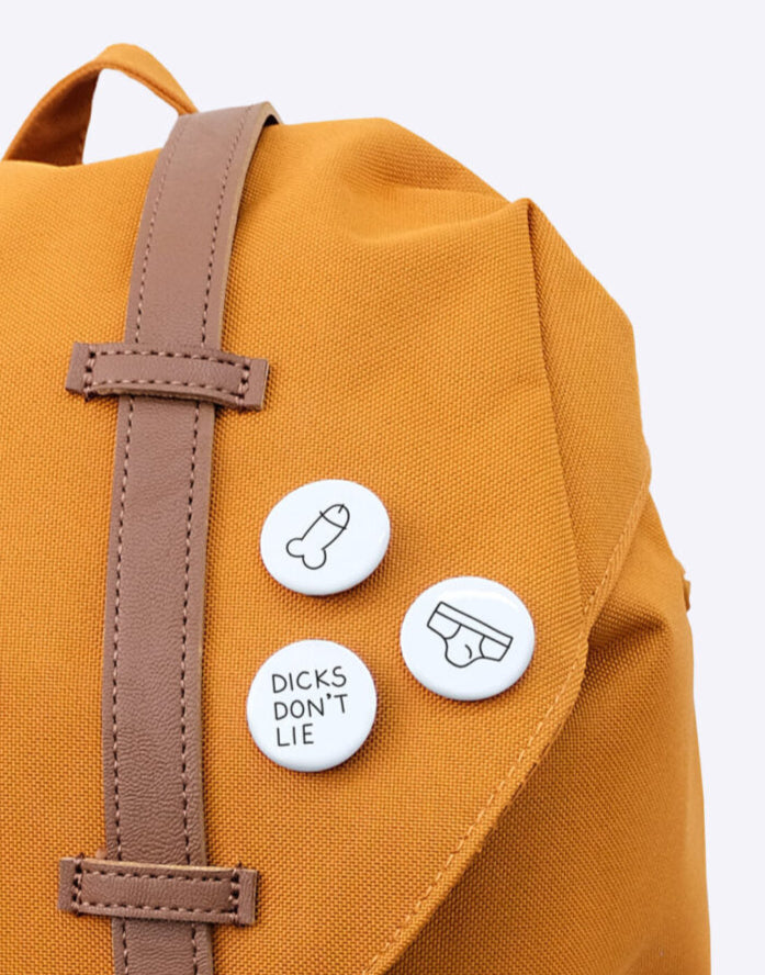 Herschel bag in terra cotta orange with 3 buttons on it. Butt-Ons from Dicks Don't Lie with line drawings on them. A drawing of a penis, a brief underwear en the logo of Dicks Don't Lie.