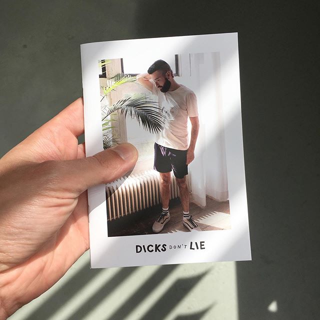 Hand holding a Dicks Don't Lie catalogue with a handsome guy on it with a beard. Wearing a short, Vans, sport socks and glasses. He is standing in front of a heater, a plant an curtains.